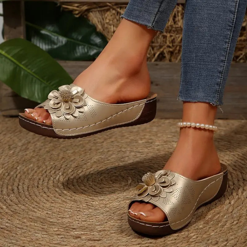 Zylia golden slip-on sandals featuring a dimensional floral embellishment and perforated strap, worn on a model's feet with blue jeans and a pearl anklet, showcasing their casual summer style.