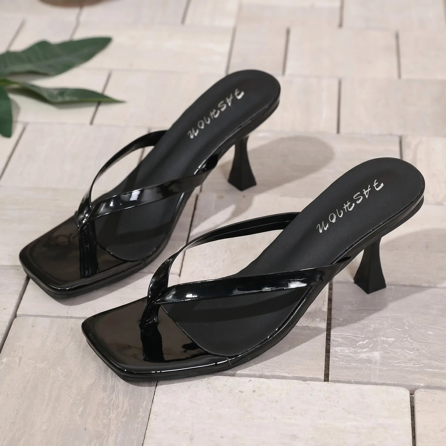 Zylira black patent square toe heeled flip flop sandals with delicate thong straps and sculptural stiletto heels, shown from a three-quarter angle on a tiled surface.