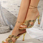Model's feet adorned with Zylith gold strappy heeled mules, featuring a square toe and a flared sculptural heel against a glamorous, sparkling background.