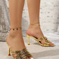 Zylith gold strappy heeled mules with a square toe, shown on a model wearing a delicate anklet, featuring a flared sculptural heel and crisscross straps against a sparkly, draped fabric background.
