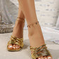 Gold strappy heeled mules with a square toe and multiple crisscross straps, worn on a woman's feet showcasing a glamorous studio setting with sparkling surfaces and draped fabric.
