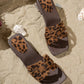 Zyloria chic summer flat slide sandals in leopard print with layered faux suede straps and a dark wood-effect stacked heel, resting on a sandy beach with shells.