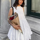 A woman models a sleeveless white tiered mini dress with a ruffled high neckline, holding a bouquet of red roses. The dress is A-line and falls above the knee, styled with dark boots in an outdoor, urban setting.