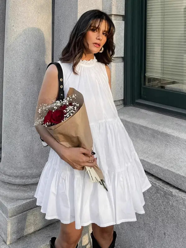 A woman models a sleeveless white tiered mini dress with a ruffled high neckline, holding a bouquet of red roses. The dress is A-line and falls above the knee, styled with dark boots in an outdoor, urban setting.
