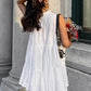Woman wearing a white sleeveless tiered mini dress with a ruffled high neckline, viewed from the back with a three-quarter angle, holding flowers outdoors.