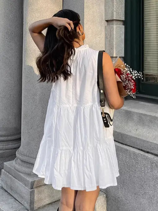 Woman wearing a white sleeveless tiered mini dress with a ruffled high neckline, viewed from the back with a three-quarter angle, holding flowers outdoors.