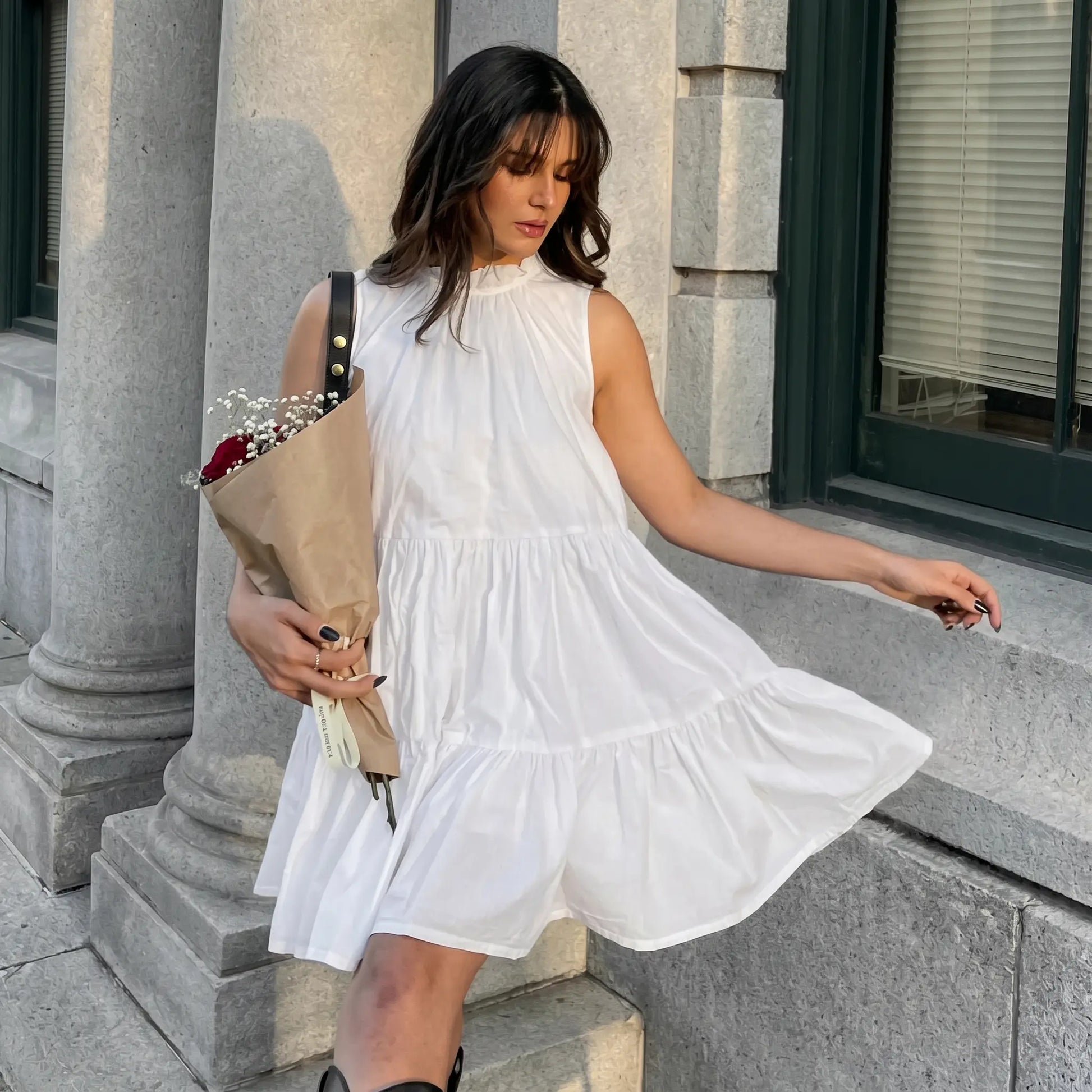 A woman in a flowing white sleeveless tiered mini dress with a ruffled high neckline, holding a bouquet of flowers, embodying romantic charm in an outdoor setting.