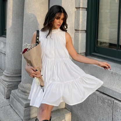 A woman in a flowing white sleeveless tiered mini dress with a ruffled high neckline, holding a bouquet of flowers, embodying romantic charm in an outdoor setting.