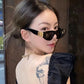 A woman models glossy black Zylos cat-eye sunglasses with gold interlocking BB logo hardware, styled with a black top and gold necklace. The sunglasses feature dark grey lenses and a bold, sculpted frame, offering a chic, designer look.