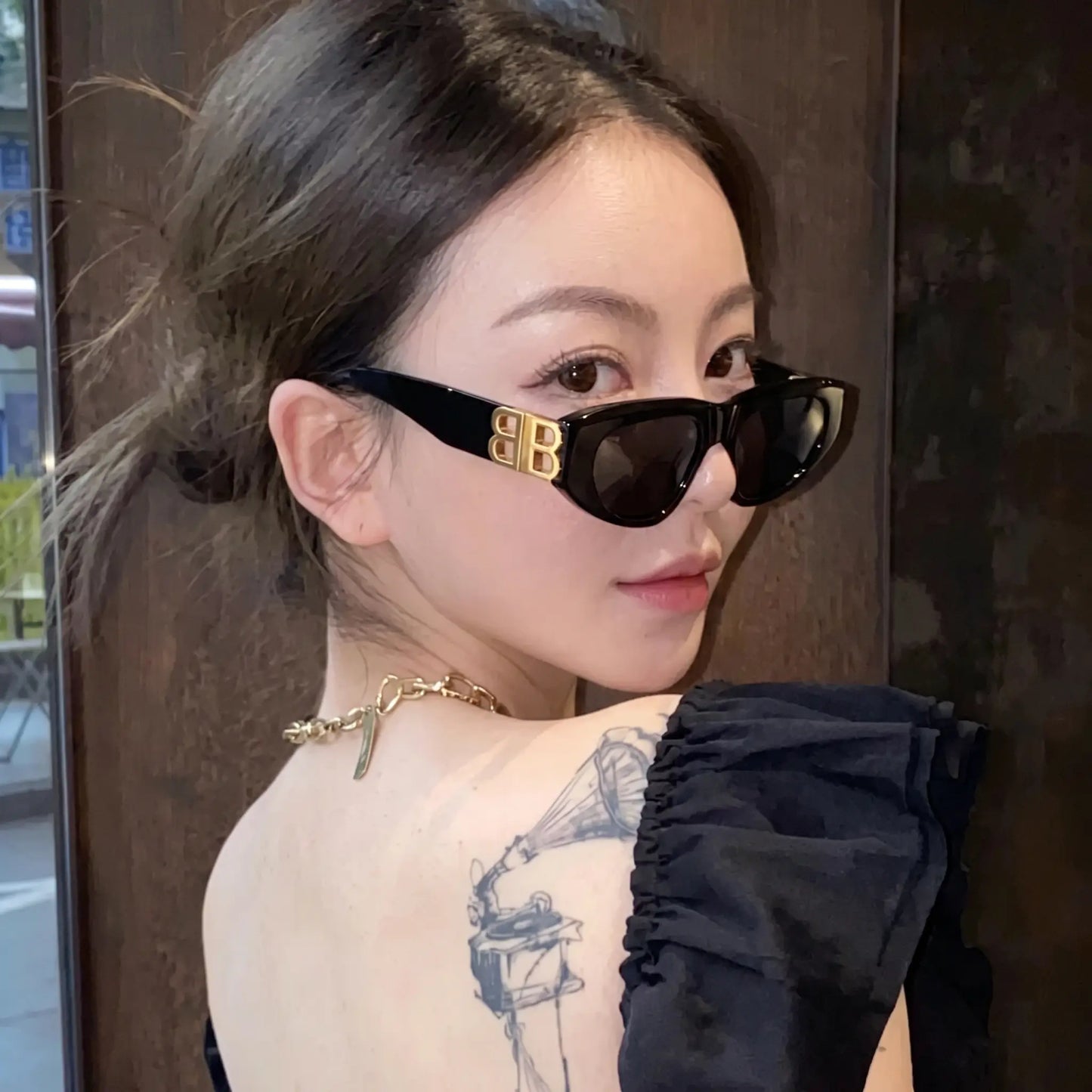 A woman models glossy black Zylos cat-eye sunglasses with gold interlocking BB logo hardware, styled with a black top and gold necklace. The sunglasses feature dark grey lenses and a bold, sculpted frame, offering a chic, designer look.