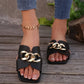 Black Zylos flat slide sandals with a chunky gold chain across the vamp, featuring a square toe and textured faux leather strap, worn by a model.
