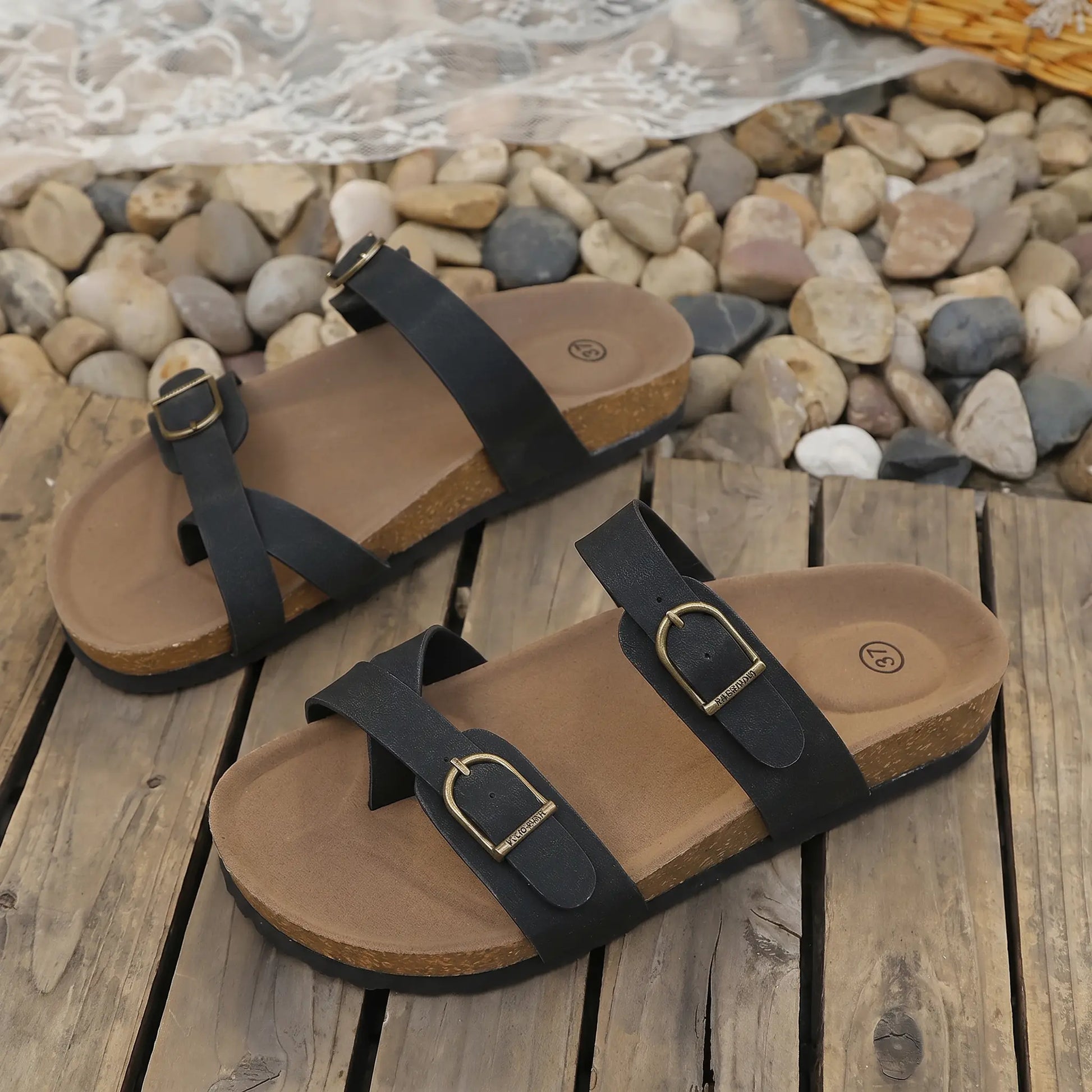 Black Zylos cork sole slip-on sandals with adjustable antique brass buckle straps, displayed on a rustic wooden surface with decorative stones.