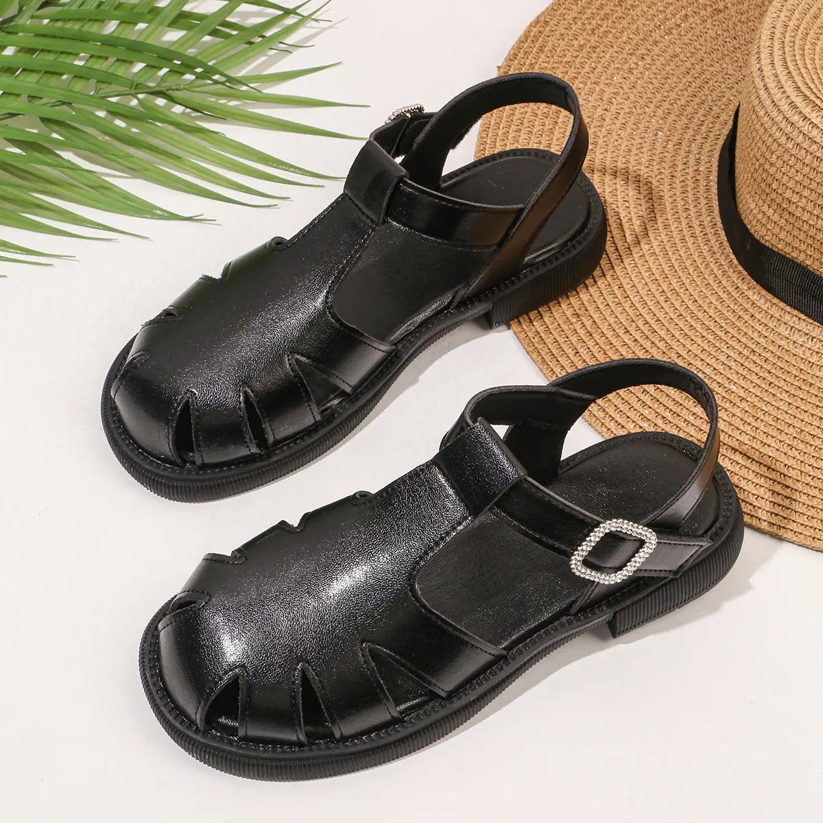 A pair of Zylos black flat sandals with T-strap design and sparkling jeweled square buckles, presented alongside a straw hat and green foliage.