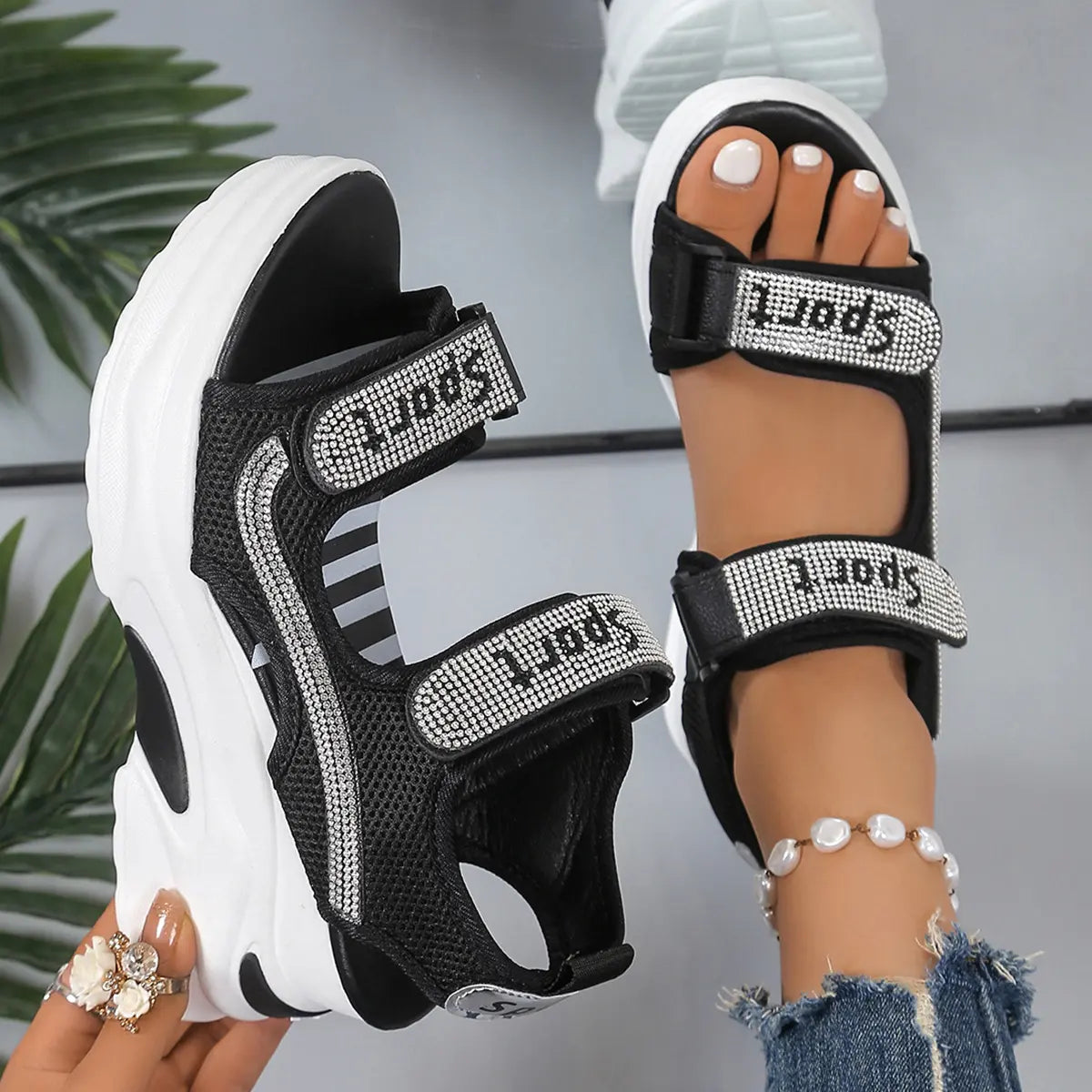 Zylos black mesh platform sandals adorned with sparkling "SPORT" rhinestone-embellished straps, shown on a foot with a pearl ankle bracelet and held by a hand showcasing a floral ring.