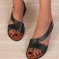 Close-up view of black Zylos Roman flat sandals on feet, showcasing the geometric faux leather straps, ankle strap, and studded sole edge detail. The open-toe design reveals a tan footbed and dark red pedicure.