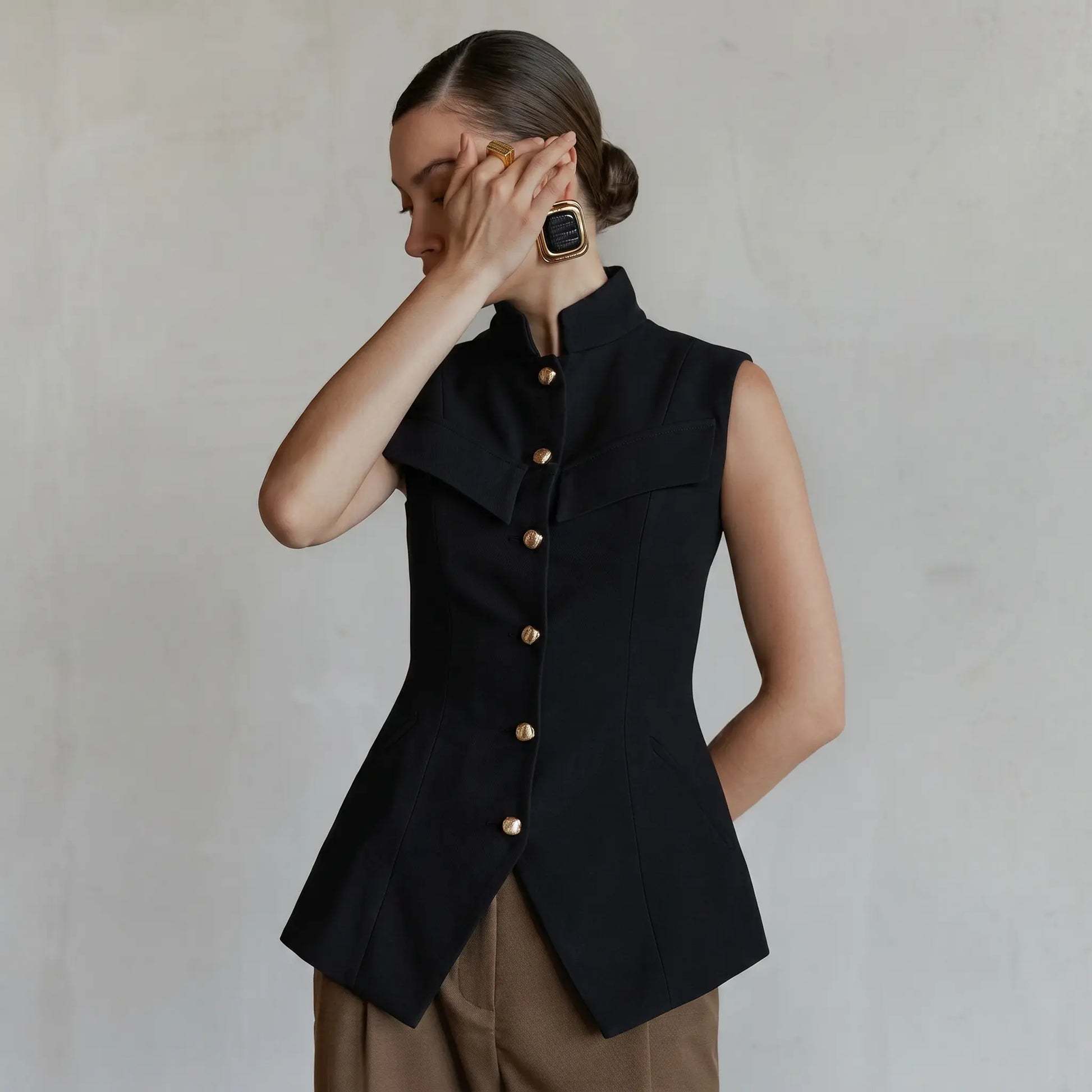 A Zylos black sleeveless tailored vest featuring a mandarin collar and gold button front, styled on a model with taupe trousers and statement earrings.