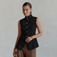 Elegant black sleeveless vest by Zylos, featuring a sharp mandarin collar, gold-tone button front, and a cinched waist with a flared peplum hem and split vent, worn by a model against a neutral studio background.
