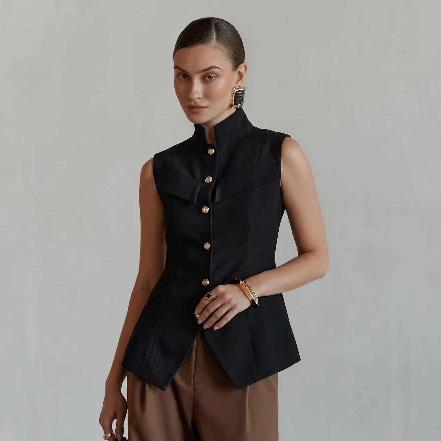 Elegant black sleeveless vest by Zylos, featuring a sharp mandarin collar, gold-tone button front, and a cinched waist with a flared peplum hem and split vent, worn by a model against a neutral studio background.
