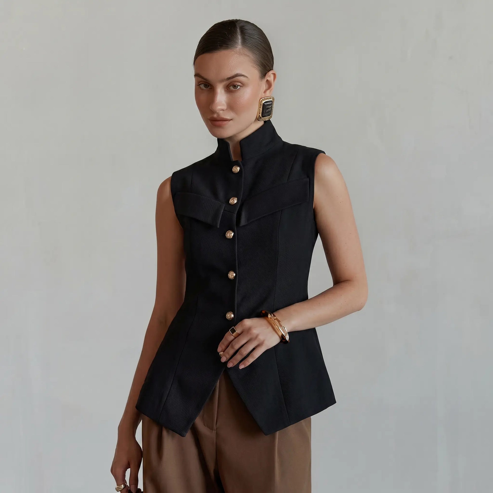 Elegant black sleeveless vest by Zylos, featuring a sharp mandarin collar, gold-tone button front, and a cinched waist with a flared peplum hem and split vent, worn by a model against a neutral studio background.