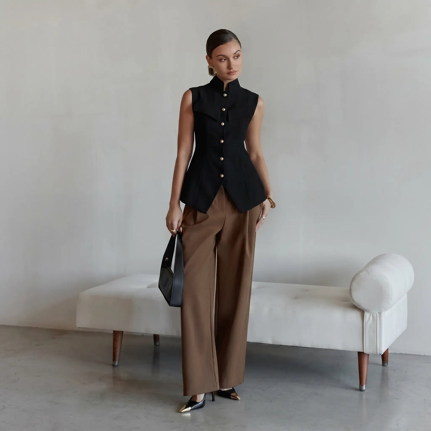 Woman wearing a tailored black sleeveless vest with a high mandarin collar and polished gold buttons, paired with taupe trousers and a black handbag.