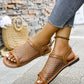 Brown mesh and faux leather flat sandals with a round toe and buckle strap, worn by a woman with white-painted toenails and grey jeans, styled with a straw bag in a relaxed outdoor setting.