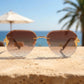 Gold-tone rimless aviator sunglasses with brown gradient lenses and intricate chain-link temple details, set against a sunny beach background with palm trees.