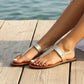 Woman wearing Zylos gold metallic textured flat sandals with an adjustable ankle strap, relaxing by the water on a wooden pier.