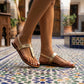 A woman's feet wearing Zylos gold metallic textured t-strap flat sandals with an adjustable ankle strap, standing on an ornate tiled patio.