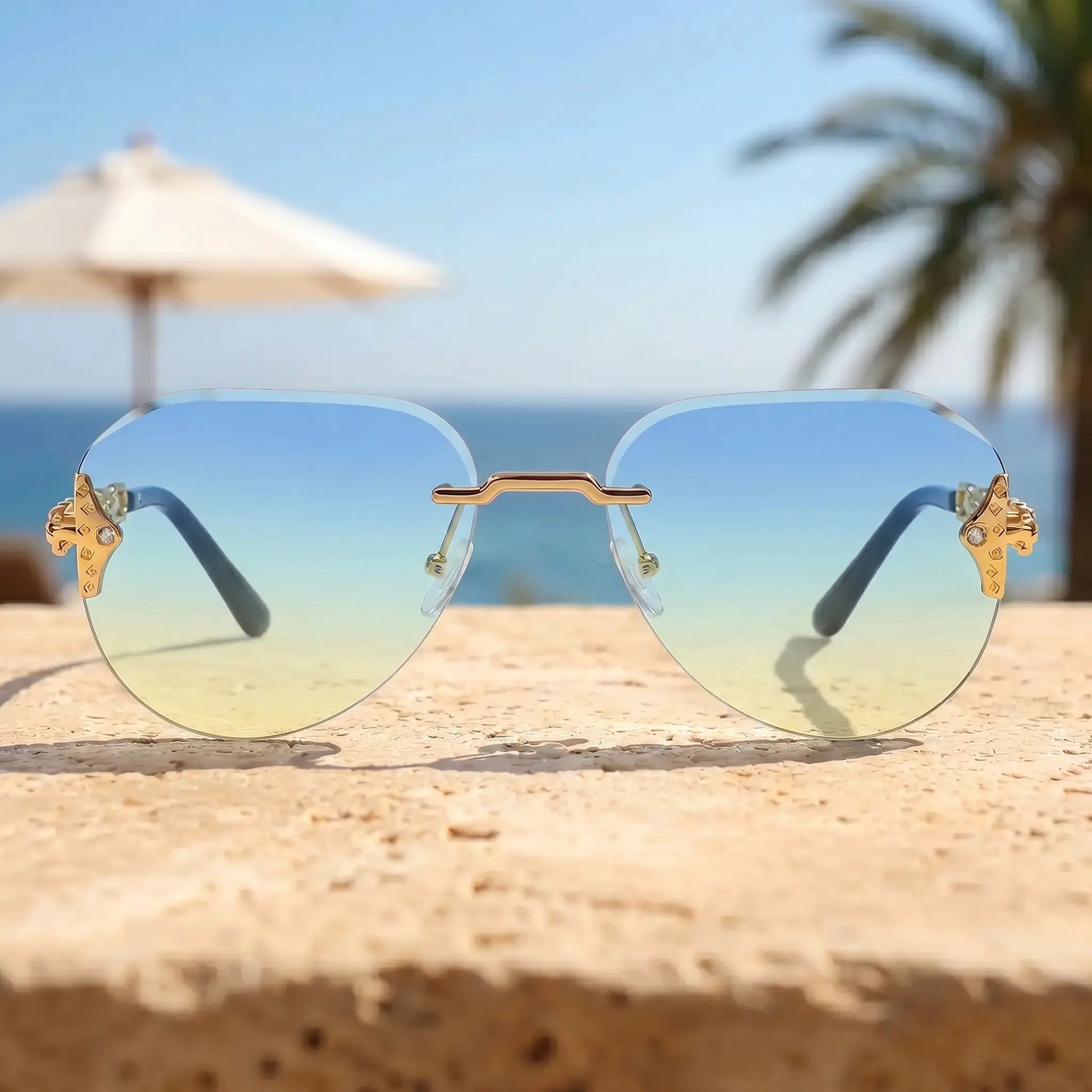 Gold-tone rimless aviator sunglasses featuring gradient blue-to-yellow lenses and ornate chain-link temple details, set against a sunny beach backdrop with ocean and palm trees.