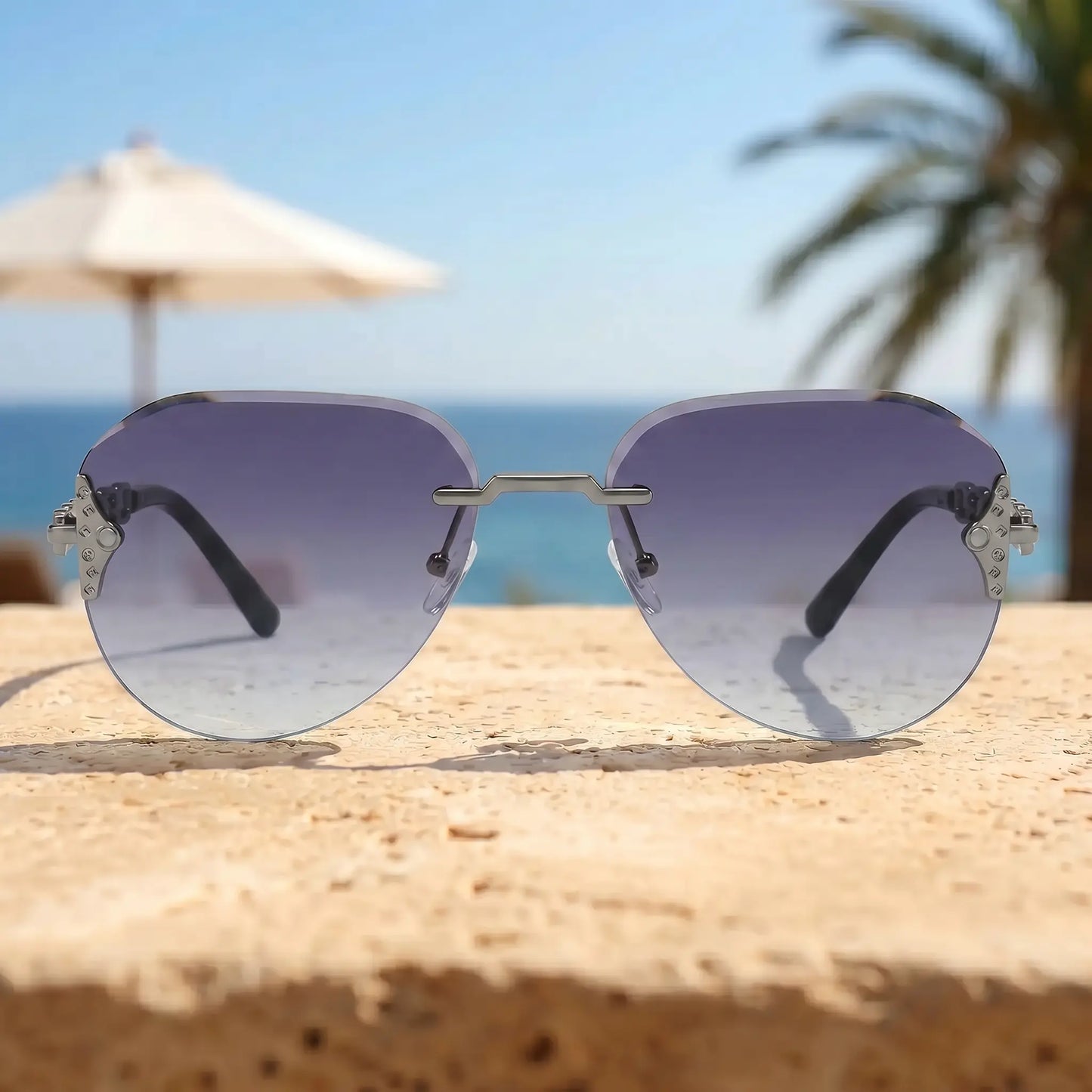Zylos gold-tone rimless aviator sunglasses with gradient grey lenses and intricate chain-link temple details, displayed on a stone surface with a beach and palm tree background. UV400 protection.
