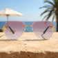 Zylos gold-tone rimless aviator sunglasses with pink grey gradient lenses, intricate chain-link temple details, showcased on a textured surface with a beach and ocean backdrop.