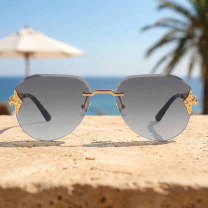 Gold-tone rimless aviator sunglasses with intricate chain-link temples and gradient grey lenses, set against a blurred beach backdrop with palm trees and an umbrella.