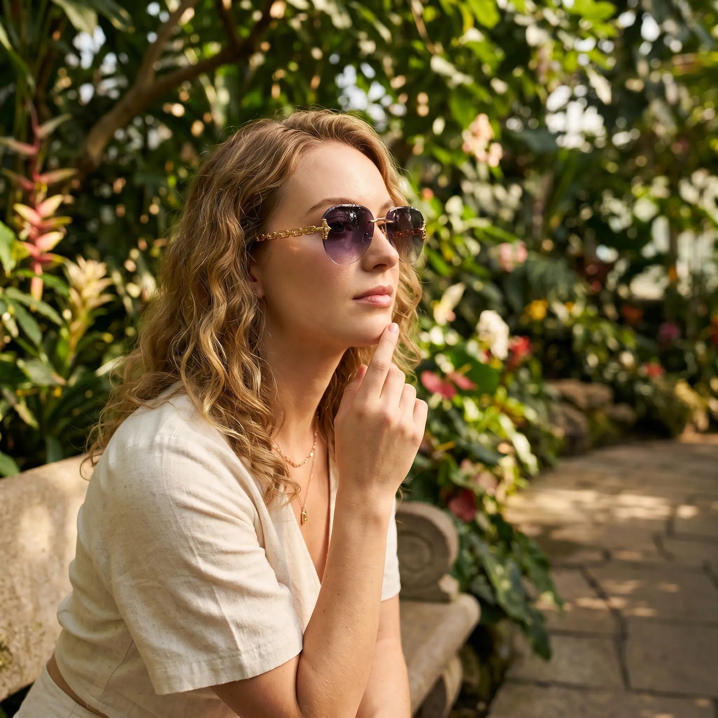 A woman wearing gold-tone rimless aviator sunglasses with chain-link temples, sitting on a bench in a lush garden setting. The gradient grey lenses offer UV400 protection, presenting a luxurious and timeless statement accessory.