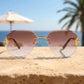 Gold-tone rimless aviator sunglasses with ornate chain-link temples and brown pink gradient lenses, styled on a textured surface with a beach and ocean backdrop.