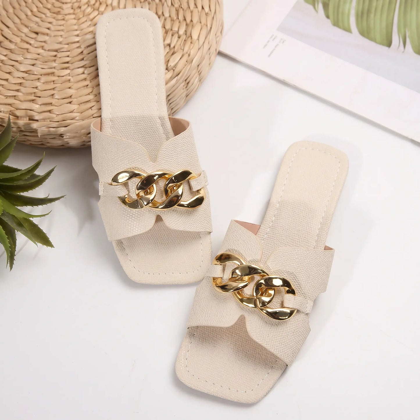 A pair of ivory Zylos flat slide sandals featuring a square toe and a wide strap adorned with a chunky gold-tone chain, presented on a studio background with subtle styling elements.