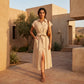 A woman models a belted midi linen shirt dress in oatmeal beige, featuring cap sleeves and a high side slit, set against an elegant desert architectural backdrop.