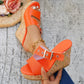 A pair of Zylos vibrant orange peep-toe wedge sandals are worn by a model, showcasing the cork-effect wedge with multi-colored flecks, adjustable silver buckle strap, and smooth PU leather. Styled with distressed denim jeans, the lifestyle image includes a straw hat with flowers in the background.