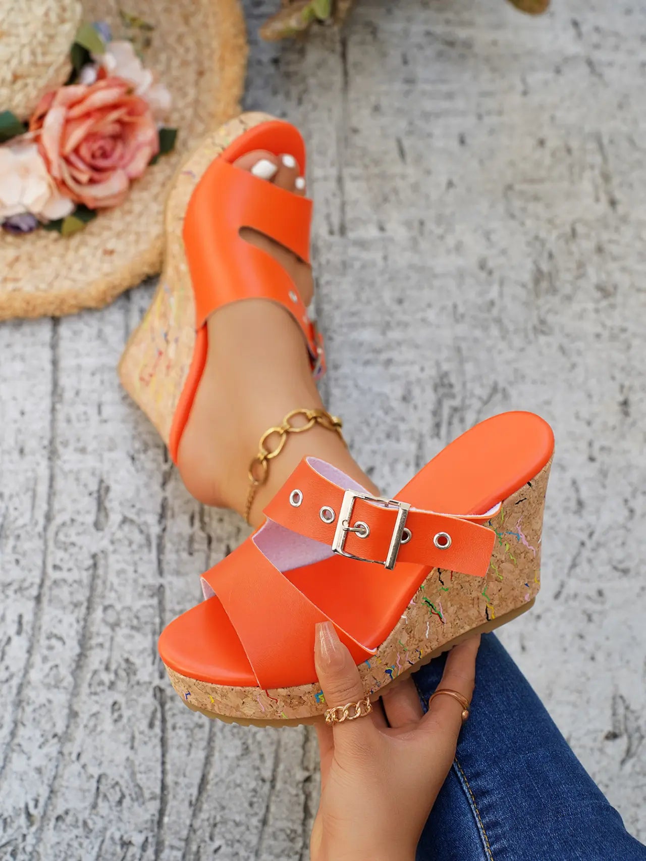 A pair of Zylos vibrant orange peep-toe wedge sandals are worn by a model, showcasing the cork-effect wedge with multi-colored flecks, adjustable silver buckle strap, and smooth PU leather. Styled with distressed denim jeans, the lifestyle image includes a straw hat with flowers in the background.