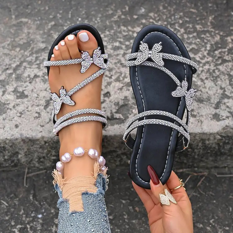 A close-up view of black Zylos rhinestone butterfly flat sandals adorned with sparkling crystal straps and butterfly embellishments, worn on a foot paired with distressed denim.
