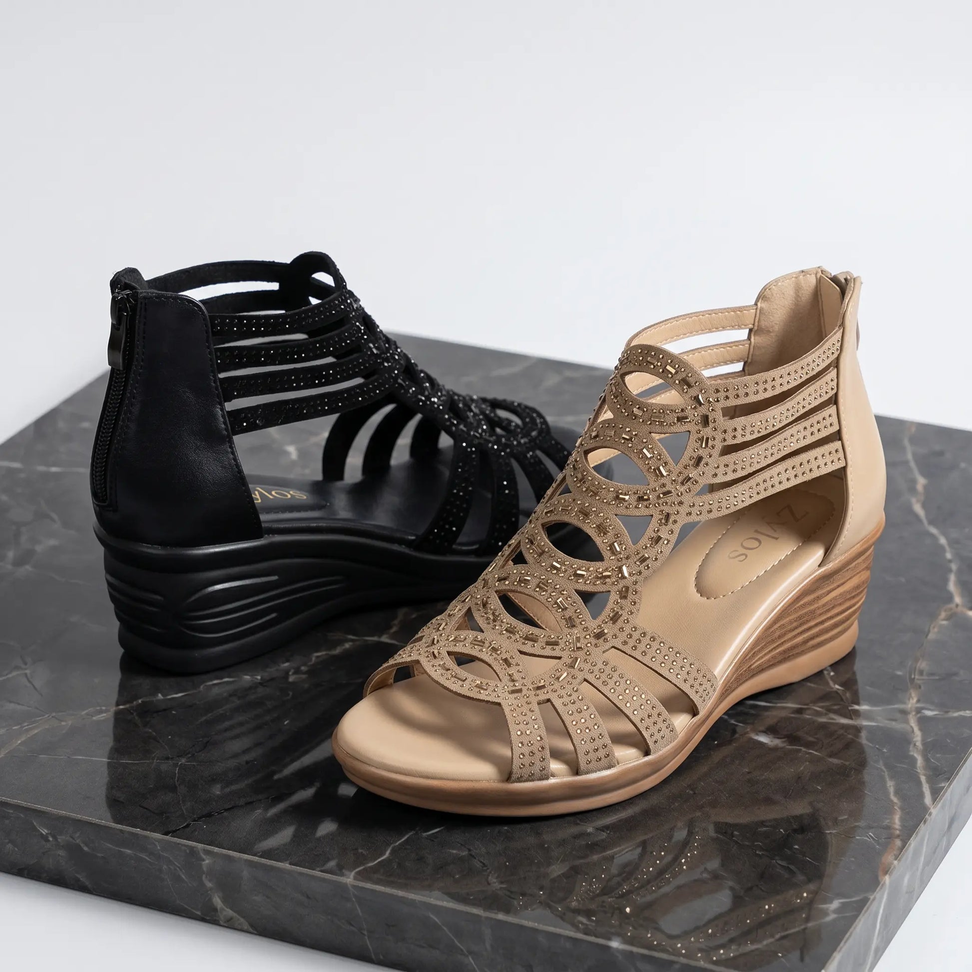 Two pairs of Zylos rhinestone wedge sandals are displayed on a marble surface: one in black and one in a golden sand khaki, showcasing their Roman-style straps and sparkling embellishments.