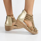 Close-up of Zylos rhinestone wedge sandals in golden sand on a model's feet, featuring intricate straps, a back zipper closure, and a gold-tone ankle chain.