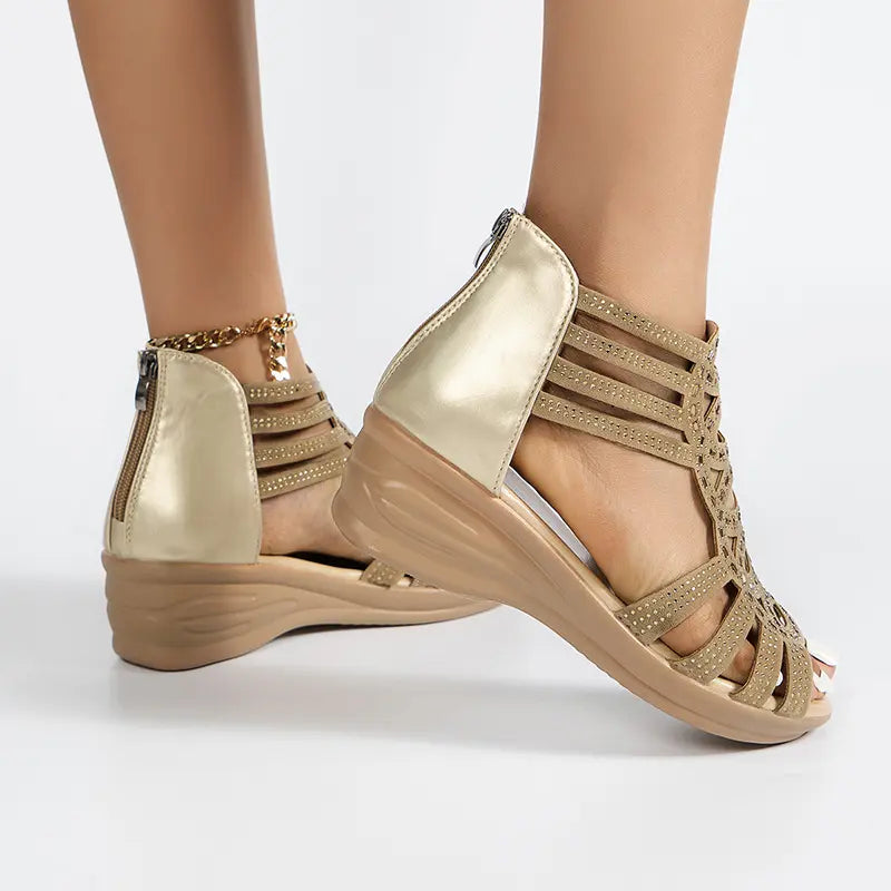 Close-up of Zylos rhinestone wedge sandals in golden sand on a model's feet, featuring intricate straps, a back zipper closure, and a gold-tone ankle chain.