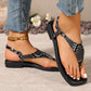 Bohemian Zylos flat sandals with elegant snake print are worn by a model with distressed denim, showcasing the square toe and adjustable ankle strap. White toenails and a gold anklet complete the look.