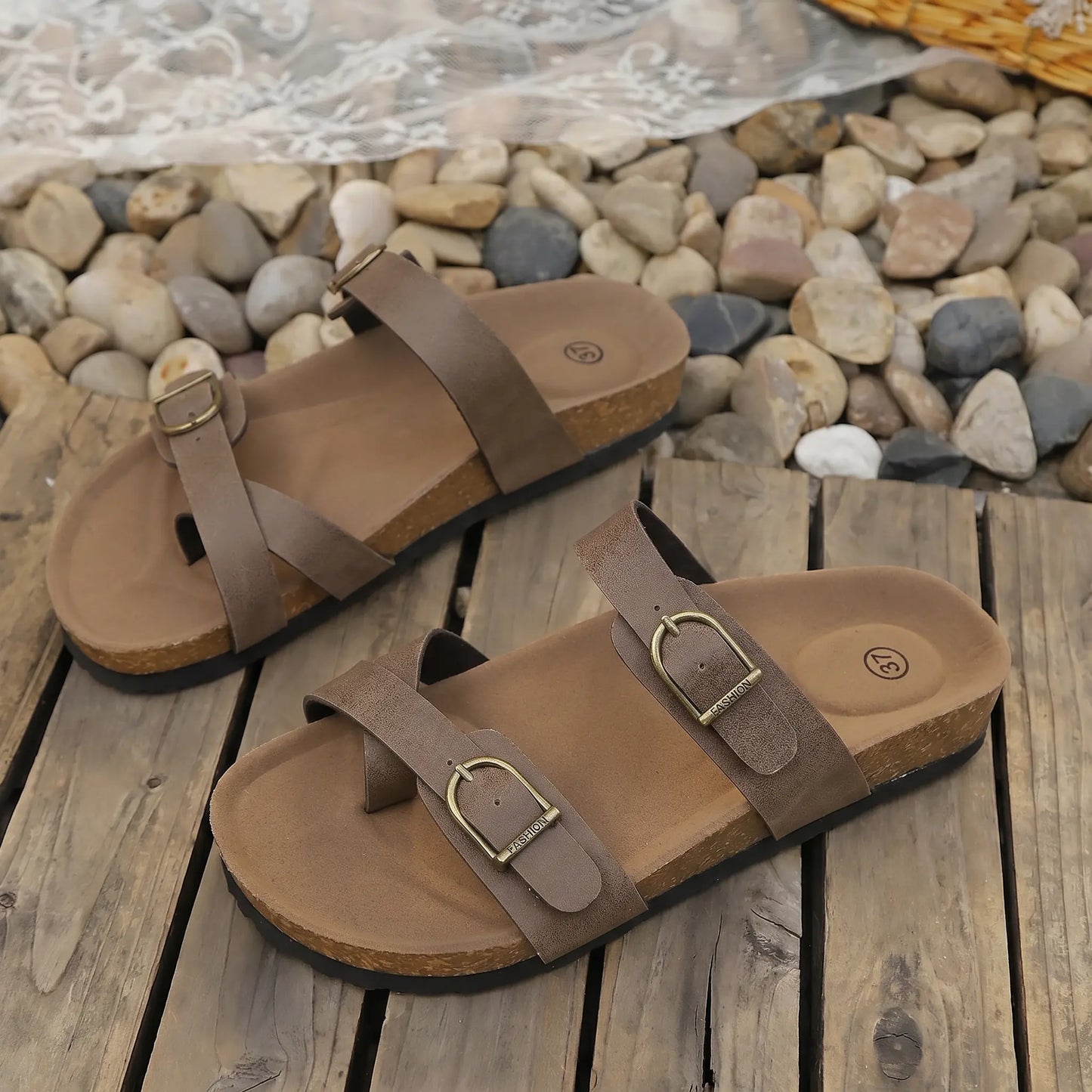 Earthy Taupe Zylos cork sole sandals with adjustable antique brass buckles, shown from a three-quarter angle on a rustic wooden and pebble background.