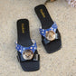 Zylpha flat slip-on sandals featuring a striking color-block design of glossy red and printed blue straps, adorned with a bold gold-tone medallion buckle with embossed detailing. Square open-toe silhouette with a black rubber sole.