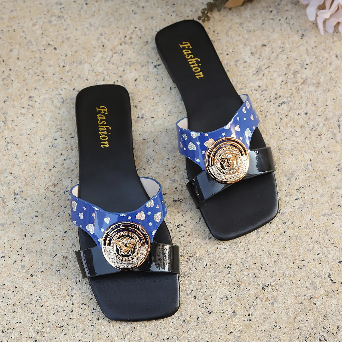 Zylpha flat slip-on sandals featuring a striking color-block design of glossy red and printed blue straps, adorned with a bold gold-tone medallion buckle with embossed detailing. Square open-toe silhouette with a black rubber sole.