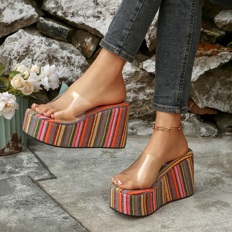Bohemian Zylpha platform wedge sandals in vibrant multi-color stripes and a clear PVC strap, shown on a woman's feet in an outdoor, casual setting with stone and plant accents.