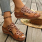 Zylphra women's chestnut brown wedge sandals featuring intricate floral laser-cut detailing on the strap, shown on a foot with an anklet, styled on a wooden deck with a woven basket.