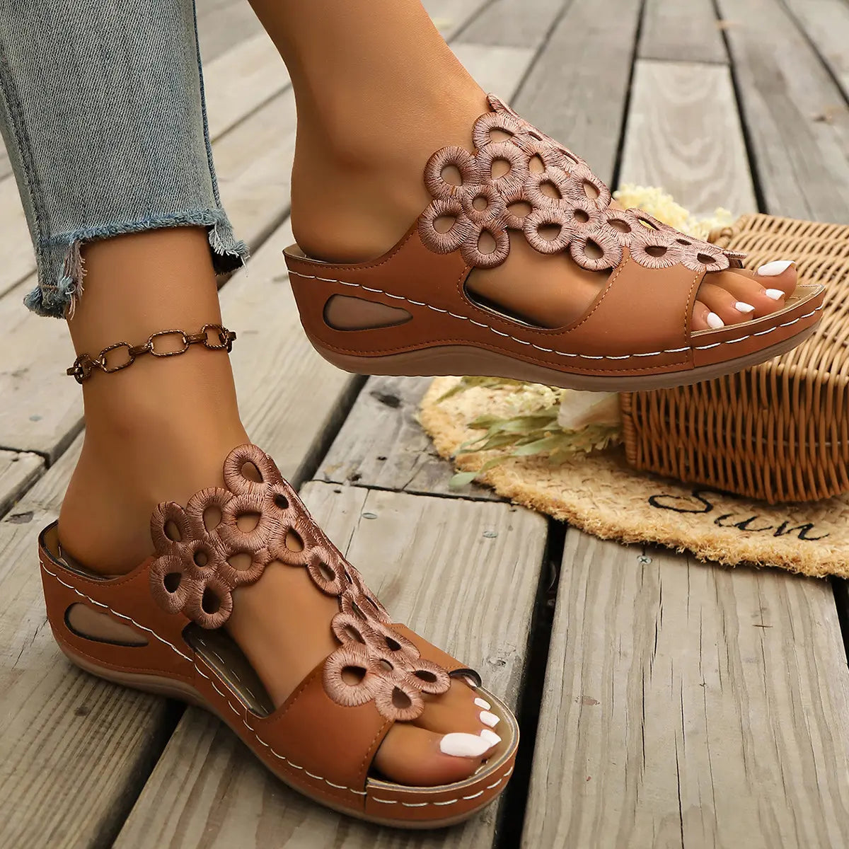 Zylphra women's chestnut brown wedge sandals featuring intricate floral laser-cut detailing on the strap, shown on a foot with an anklet, styled on a wooden deck with a woven basket.