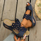 Navy blue Zylphra women's wedge sandals with intricate laser-cut floral details on the strap, shown worn on a foot with a straw hat and greenery in the background.